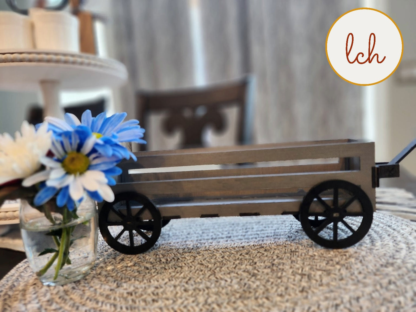 Wooden wagon insert with black wheels displayed on a woven mat beside a vase with blue and white flowers